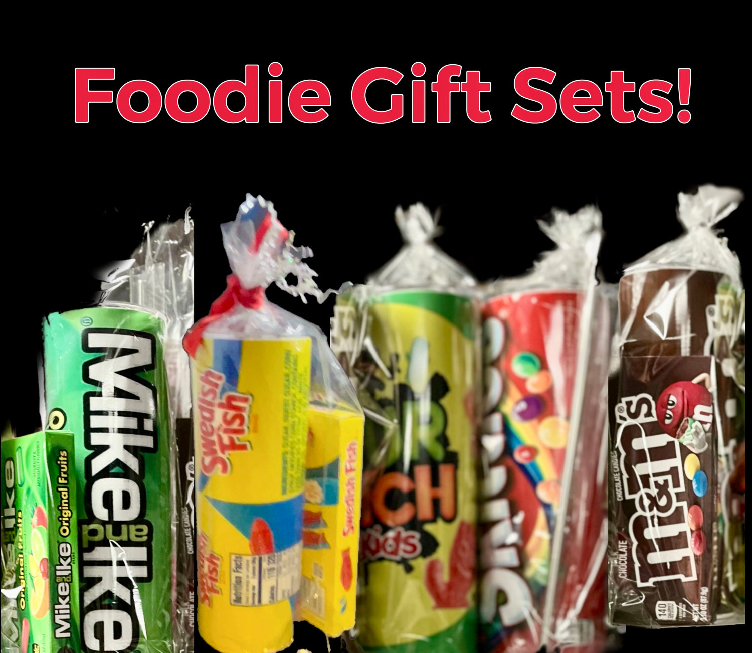 A selection of wrapped candy gift sets against a black background. Visible candies include Mike and Ike, Swedish Fish, Airheads, Skittles, and M&M's. The text "Foodie Gift Sets!" is in red at the top.