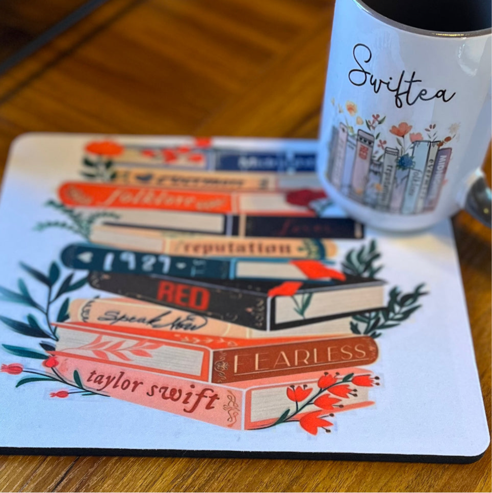 A charming Swiftea Gift Set by Kreative Kreationz sits on a table, featuring a white mug with the word "Swiftea" and a mousepad adorned with an illustration of book spines named after Taylor Swift albums: "Folklore," "Reputation," "1989," "Red," "Speak Now," and "Fearless." Ideal for tea enthusiasts, this delightful set combines elegance and fandom beautifully.