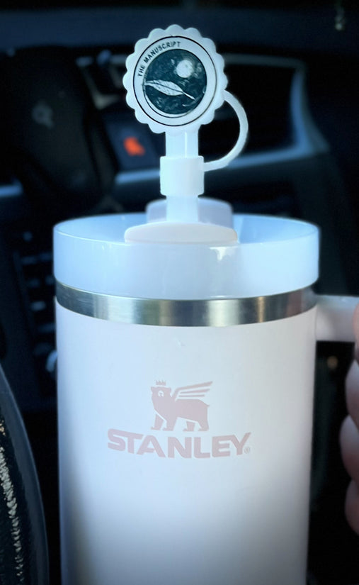 Stanley cup with a white straw cap that’s circular on the circular cap it says “the manuscript”  with a feather in dark writing 