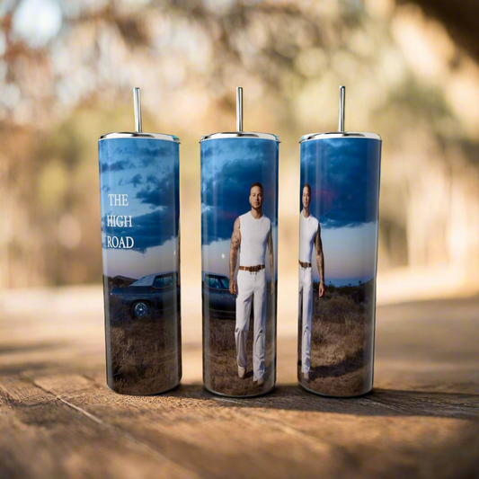 Three Kane Brown The High Road 20oz Tumblers by Kreative Kreationz are displayed outdoors, featuring lids and straws. They depict a man in a white outfit, a classic car, and "THE HIGH ROAD" text against a sky backdrop, with the blurred natural setting enhancing these eco-friendly reusable pieces.