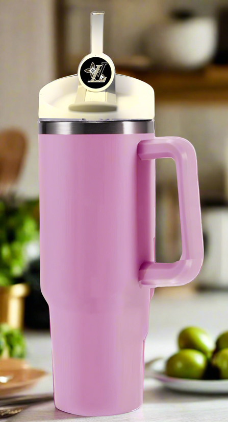 A pink insulated tumbler with a lid and Stanley tumbler accessory adorns the kitchen counter, its straw showcasing the Kreative Kreationz LV Straw Topper. In the background, blurred plants and small green fruits bring a natural touch to the scene.