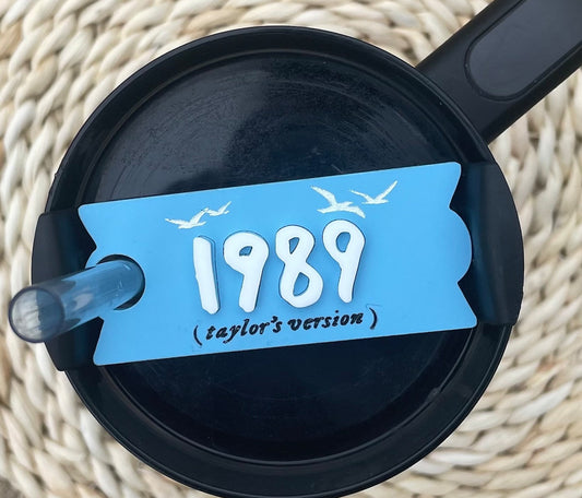 A clear, round plastic lid from a top-down view, featuring a gray sliding cover with a small circular opening on one side. The sliding cover has a logo of a winged lion wearing a crown. This customizable Kreative Kreationz Taylor Swift 1989 Taylor’s Version 20, 30, 40 ounce Stanley Lid Plate/Topper is perfect for adding flair to your drinkware collection.