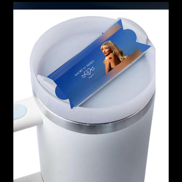 A white mug with a light blue button feature, covered by a lid. On the lid sits a blue, rectangular packaging featuring an image of a woman with blonde hair in a black outfit. The text on the packaging reads "SHOW 'N' SWEET." Crafted from premium materials, it resembles the Sabrina Carpenter Short & Sweet Stanley Lid Plate/Topper by Kreative Kreationz in quality.