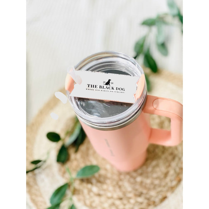 A pink tumbler with a TTPD Black Dog Stanley Lid Plate/Topper from Kreative Kreationz sits on a woven mat. On top of the lid, there is a white sticker with black text reading "THE BLACK DOG," featuring a dog silhouette and additional smaller text. Green leaves are blurred in the background.