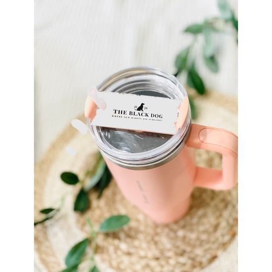 A pink tumbler with a TTPD Black Dog Stanley Lid Plate/Topper from Kreative Kreationz sits on a woven mat. On top of the lid, there is a white sticker with black text reading 