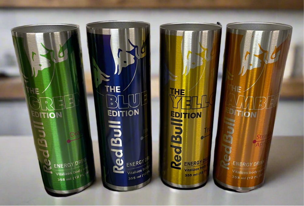 Four Red Bull energy drink cans are lined up side by side, labeled as The Green Edition (Dragon Fruit), The Blue Edition (Blueberry), The Yellow Edition (Tropical), and The Amber Edition (Strawberry-Apricot). Accompanying the colorful array is a stylish Red Bull Stainless Steel 20oz Tumbler by Kreative Kreationz.