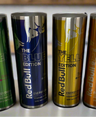 Four tall cans of Red Bull editions in green, blue, yellow, and amber colors are lined up on a table. Beside them is a sleek RB Energy Silver Tumbler by Kreative Kreationz, with 20 oz capacity—ideal for keeping your favorite edition ice-cold and ready to energize.