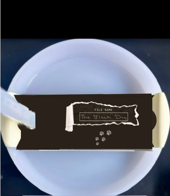 A white mug with a light blue handle is shown against a plain background. On top of the Kreative Kreationz Black Dog lid Plate/Topper, which fits 20, 30, and 40 oz Stanley cups, is a partially unwrapped chocolate bar with a black wrapper.