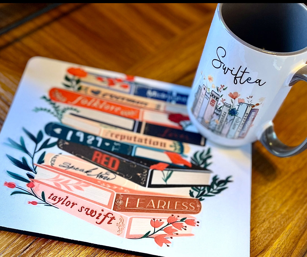 A charming Swiftea Gift Set by Kreative Kreationz sits on a table, featuring a white mug with the word "Swiftea" and a mousepad adorned with an illustration of book spines named after Taylor Swift albums: "Folklore," "Reputation," "1989," "Red," "Speak Now," and "Fearless." Ideal for tea enthusiasts, this delightful set combines elegance and fandom beautifully.