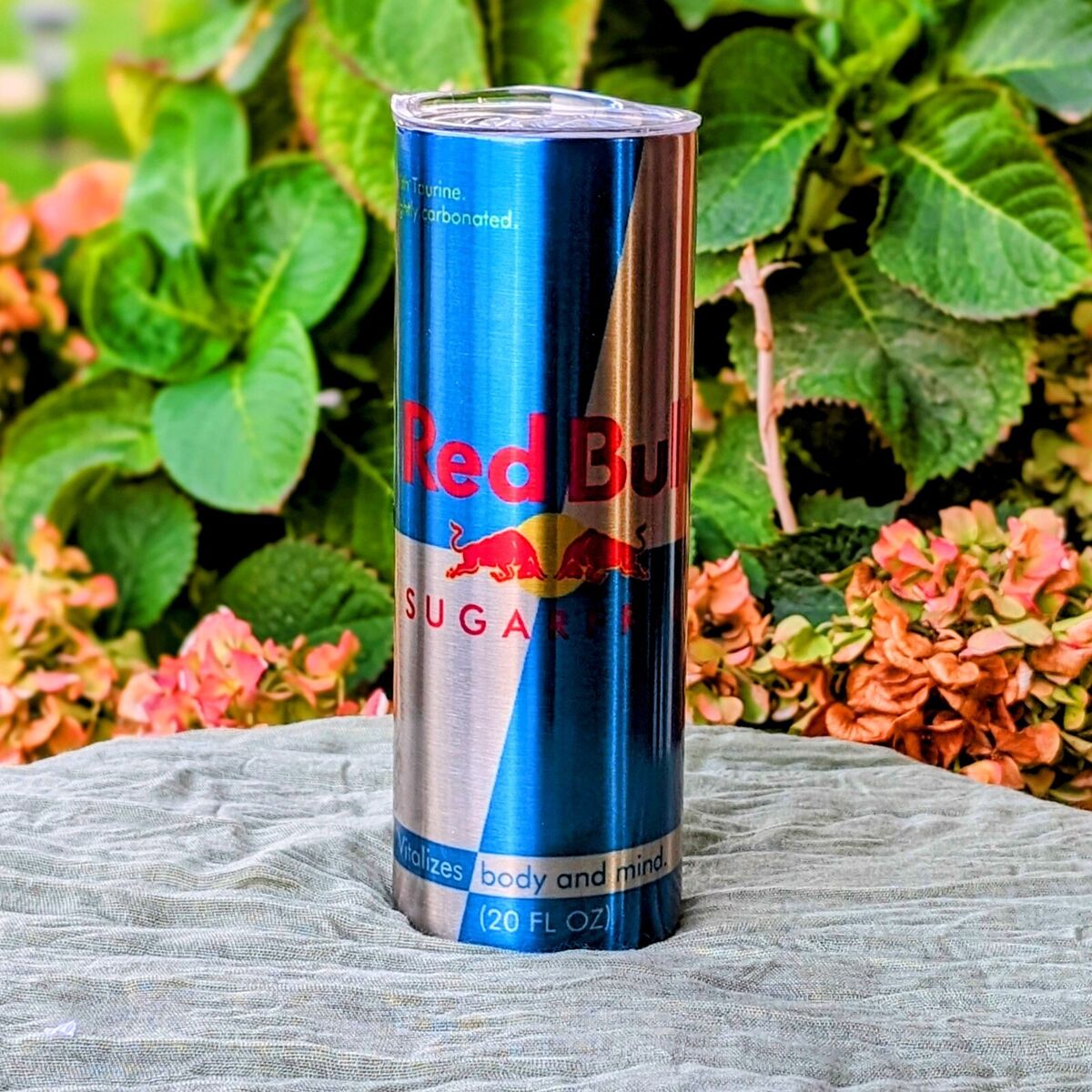 A tall, blue can of Red Bull Sugar Free rests beside a Kreative Kreationz Silver Stainless Steel 20oz Tumbler on a light green fabric surface. In the background, green leaves and pink flowers infuse a natural touch into this vibrant scene.