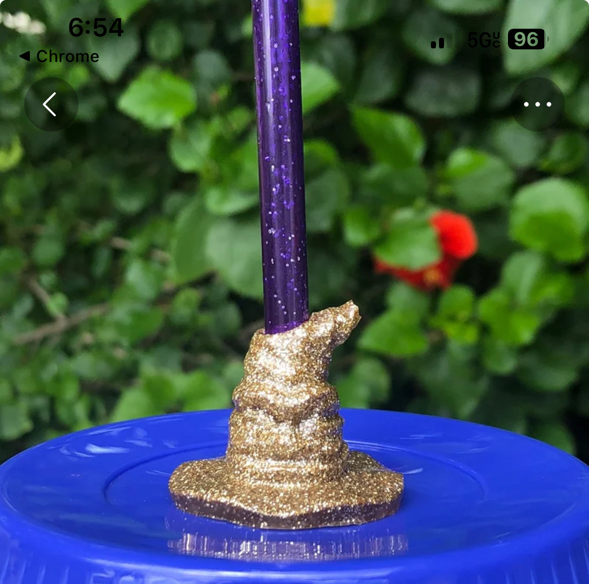 A Harry Potter Wizard | Witch Hat straw decoration from Kreative Kreationz stands upright, securely held in place by a mound of golden, glittery clay on a blue plastic surface. The background features lush green foliage and a red flower. The timestamp and battery indicator suggest the photo was taken on a mobile phone.