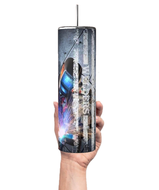 A hand holds a Welder 20oz Tumbler from Kreative Kreationz, featuring a metallic design and an image of a welder against a distressed American flag. This tall, cylindrical tumbler is part of the Welders Collection and includes a metal straw protruding from the lid.