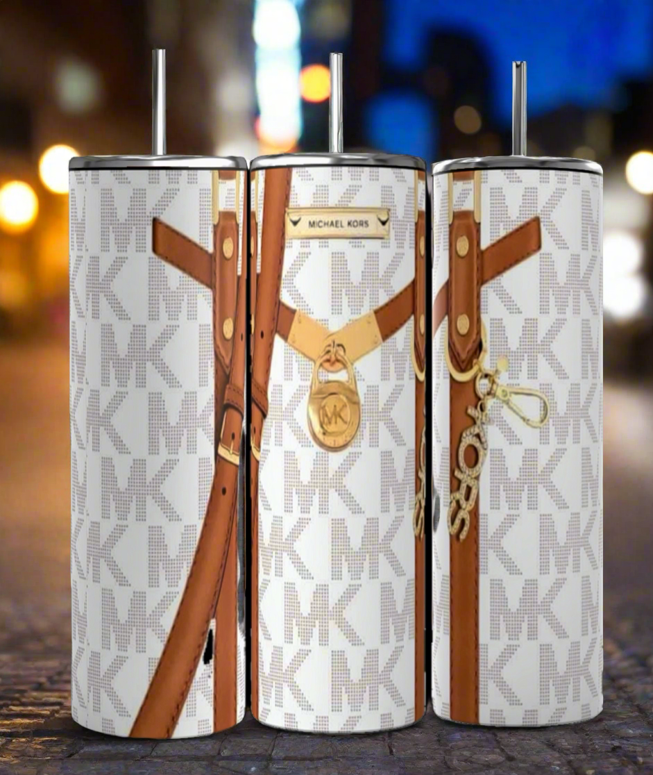 Three tall, cylindrical Kreative Kreationz Michael Kors Design Inspired White and Brown 20oz Tumblers, featuring brown strap designs and repeating "MK" initials in the background. These durable tumblers come with metal straws and keep beverages hot or cold, set against a blurred city lights background.