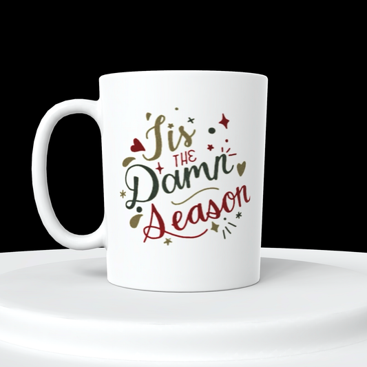 The "Tis The Damn Season Taylor Swift Ceramic Mug" by Kreative Kreationz is showcased on a white circular platform against a black background, featuring festive red, green, and gold text that reads "Tis The Damn Season." Small decorative elements like stars and hearts surround the text, making it perfect for any Taylor Swift fan.