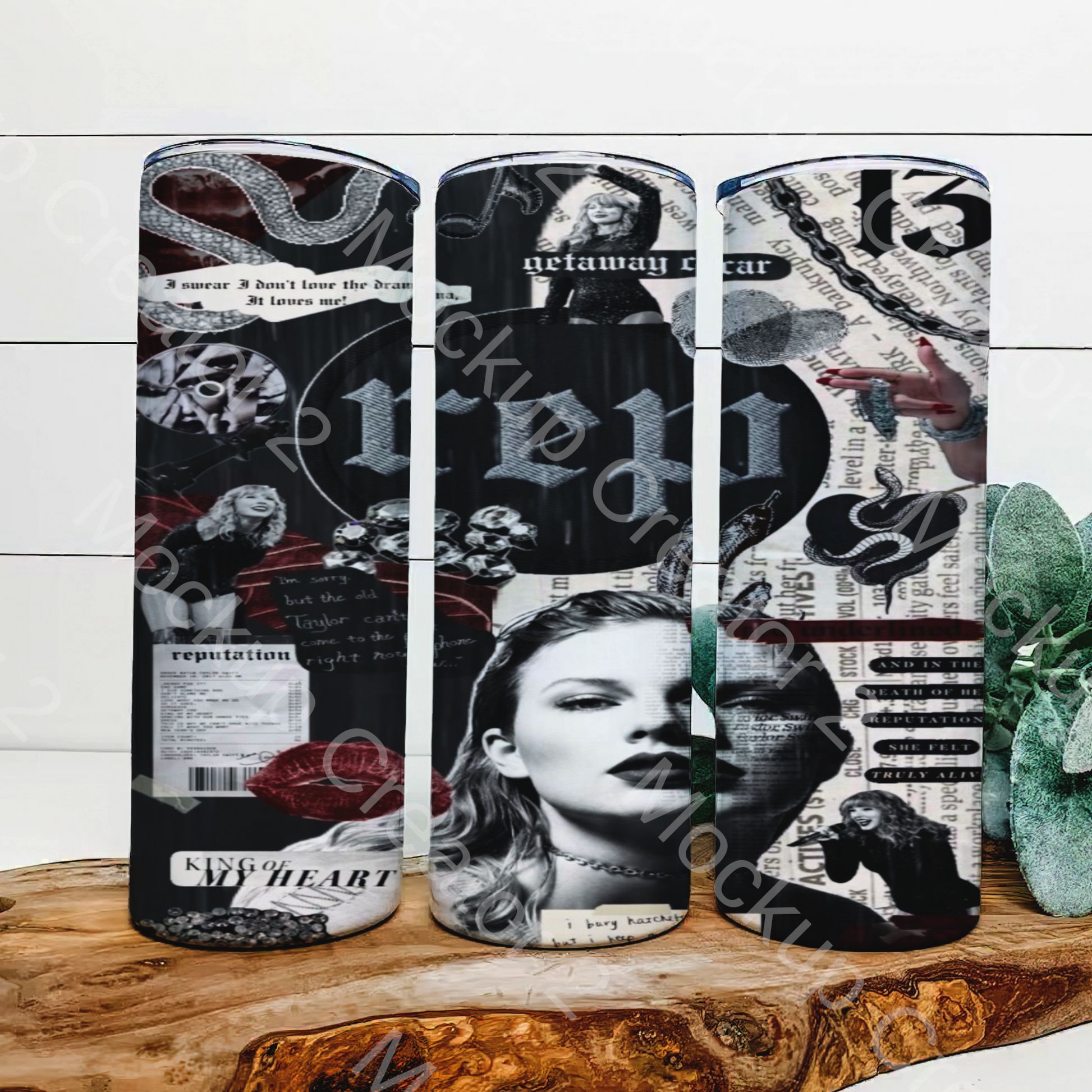 Three snowboards with black and white collage designs of newspaper clippings, gothic fonts, and portraits are displayed vertically. Nearby is a Kreative Kreationz "Rep Collage | Taylor Swift 20oz Tumbler" with a spill-proof lid and reusable straw, partially obscured by green plants.