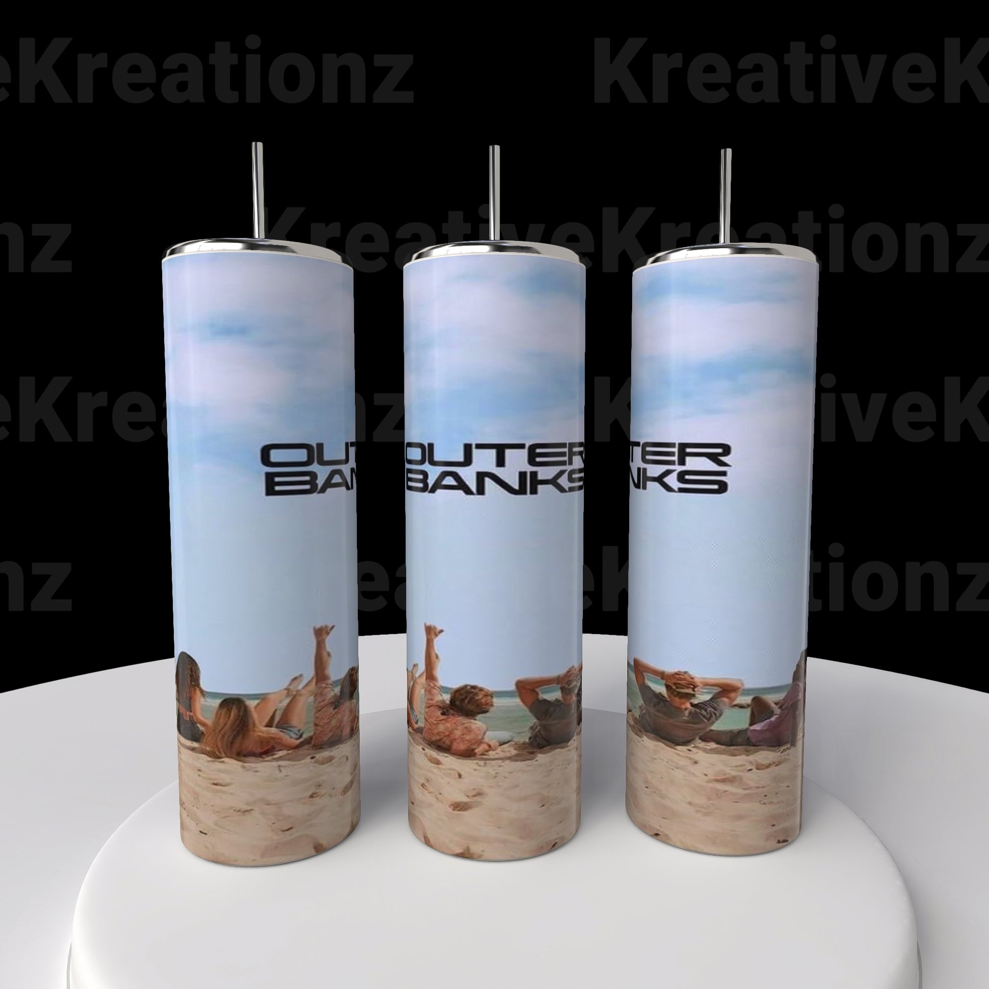 Three Outer Banks OBX 20oz Tumblers by Kreative Kreationz, tall and cylindrical, rest on a round white platform. Displaying a beach scene with "OUTER BANKS" text against a black background, each includes a spill-proof lid and metal straw.