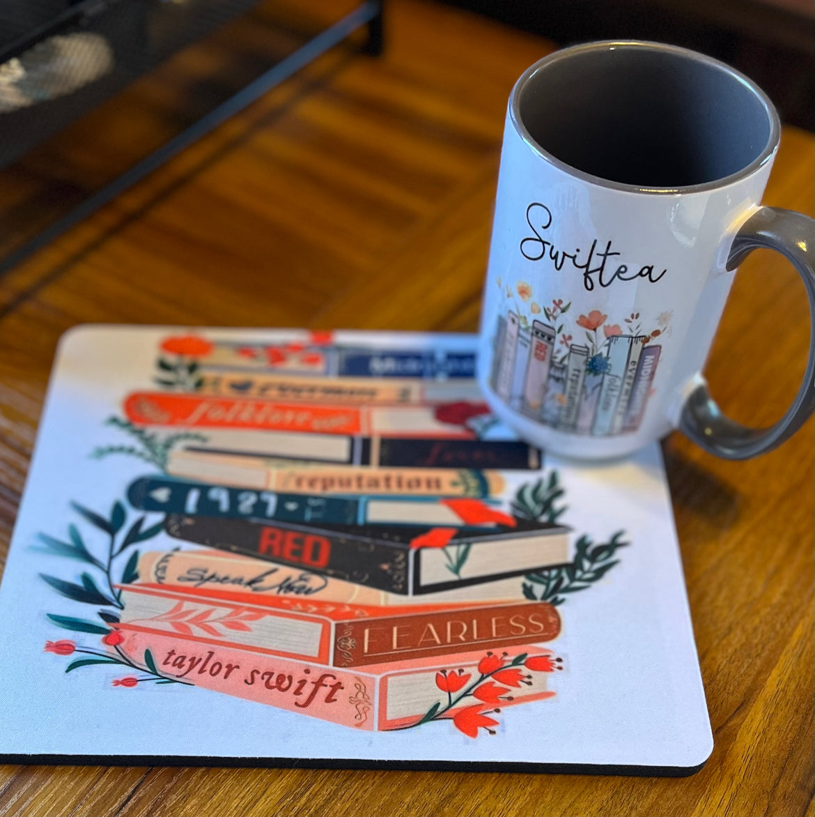 A charming Swiftea Gift Set by Kreative Kreationz sits on a table, featuring a white mug with the word "Swiftea" and a mousepad adorned with an illustration of book spines named after Taylor Swift albums: "Folklore," "Reputation," "1989," "Red," "Speak Now," and "Fearless." Ideal for tea enthusiasts, this delightful set combines elegance and fandom beautifully.