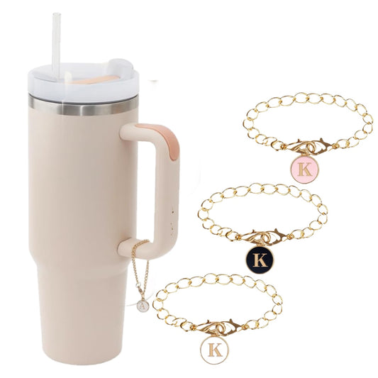 A tall, beige tumbler with a handle and straw, featuring a pink lid detail from Kreative Kreationz, is positioned next to three gold bracelet chains with round, personalized Monogram Charm Dangle pendants. The pendants display the letter "K" in light pink, black, and cream. Additionally, a sleek silver chain with a custom cup pendant catches the eye.