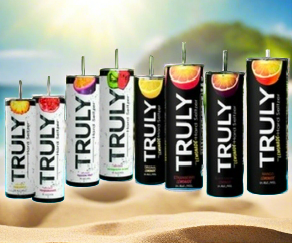 Several cans of Truly spiked & sparkling water sit on a sandy beach with the ocean and a rocky hill in the blurry background. The cans, featuring flavors like pineapple, watermelon, wild berry, and lemon, gleam brightly under the sun. A nearby 20oz tumbler from Kreative Kreationz with an add-on 3D fruit and ice lid topper adds to the summery vibe.