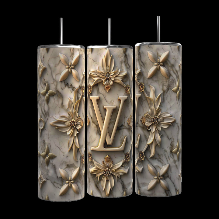 Three intricately decorated 20 oz Louis Vuitton Silver Gold tumblers by Kreative Kreationz, featuring a marbled texture and golden floral designs, stand side by side on a cobblestone street. The middle tumbler prominently showcases the initials "LV" and comes with a reusable straw. In the background, an urban setting with blurred buildings and greenery is visible.