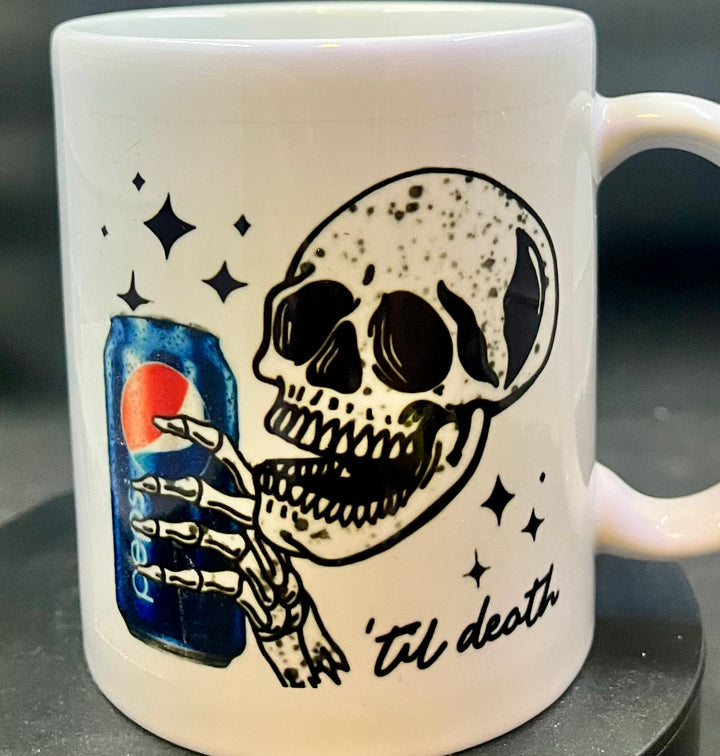 The Pepsi to death Skull Ceramic Mug by Kreative Kreationz features a striking black and white illustration of a grinning skull holding a Pepsi can. Below the image, the phrase "'til death" is inscribed, surrounded by small black stars that bring a stylish Swiftie touch for any Taylor Swift enthusiast.