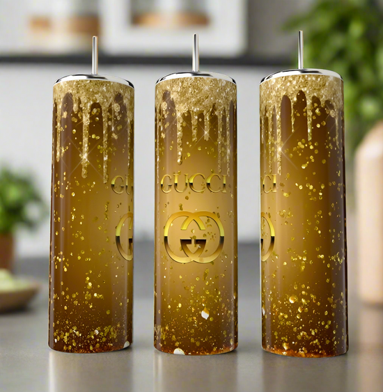 Three Gucci Gold 20oz Skinny Tumblers by Kreative Kreationz stand tall on a countertop. Filled with golden glitter and displaying the Gucci logo, they are set against a blurred backdrop of lush foliage, reflecting the sophistication of an elegant design.