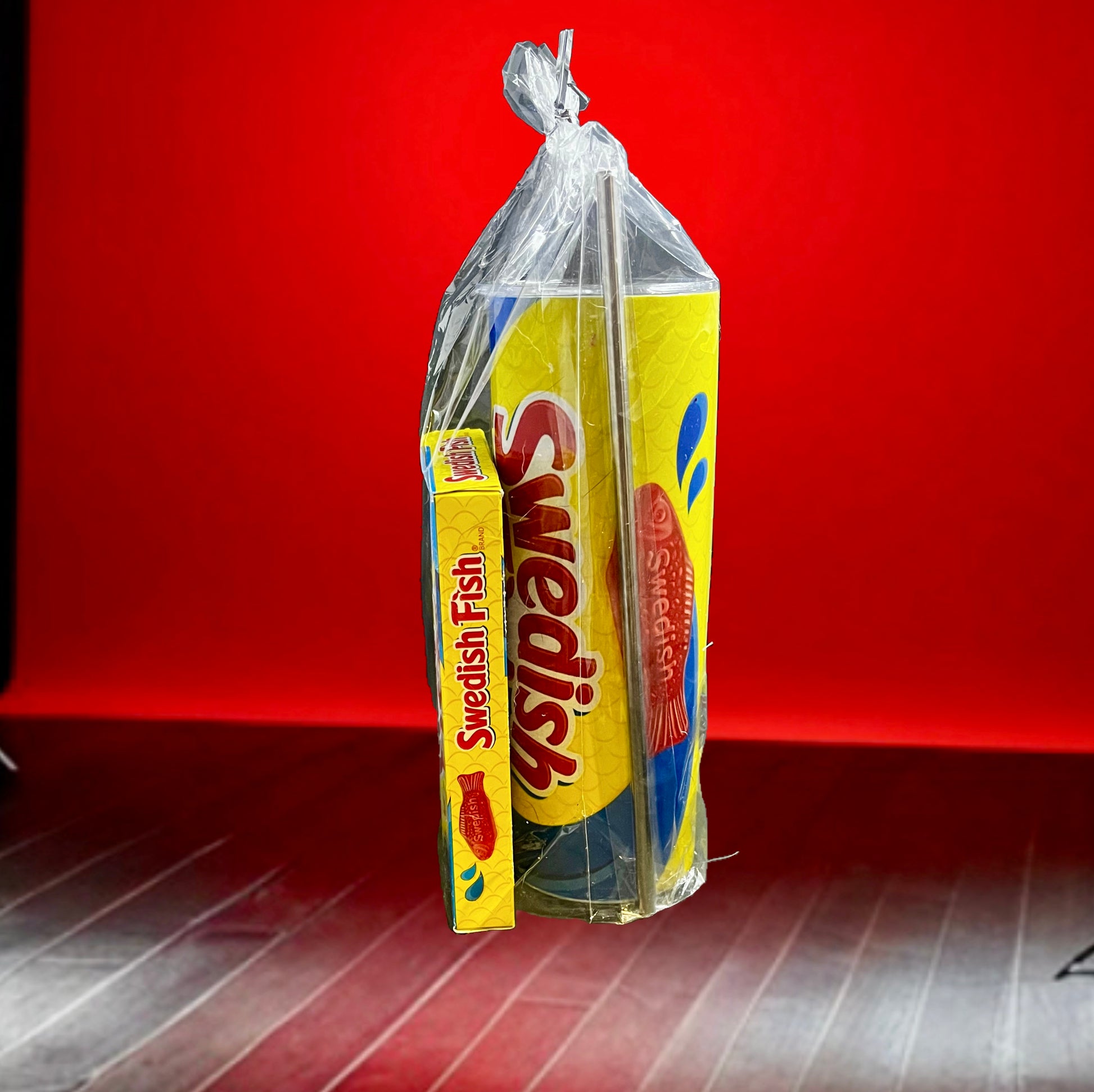 A plastic bag containing a Swedish Fish Gift Set with a 20oz stainless steel skinny tumbler, featuring yellow and blue packaging, branded "Swedish," with an illustration of a red fish. Next to it, a smaller box labeled "Swedish Fish" sits on the floor. The background is red with wooden flooring.