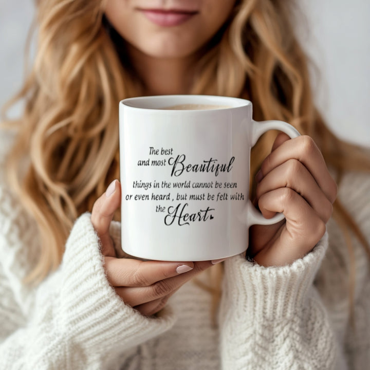 A person with long, wavy hair holds a Kreative Kreationz Personalized Couples Ceramic Mug with the quote, "The best and most beautiful things in the world cannot be seen or even heard, but must be felt with the heart," wearing a cozy white sweater—perfect for gifting to loved ones.