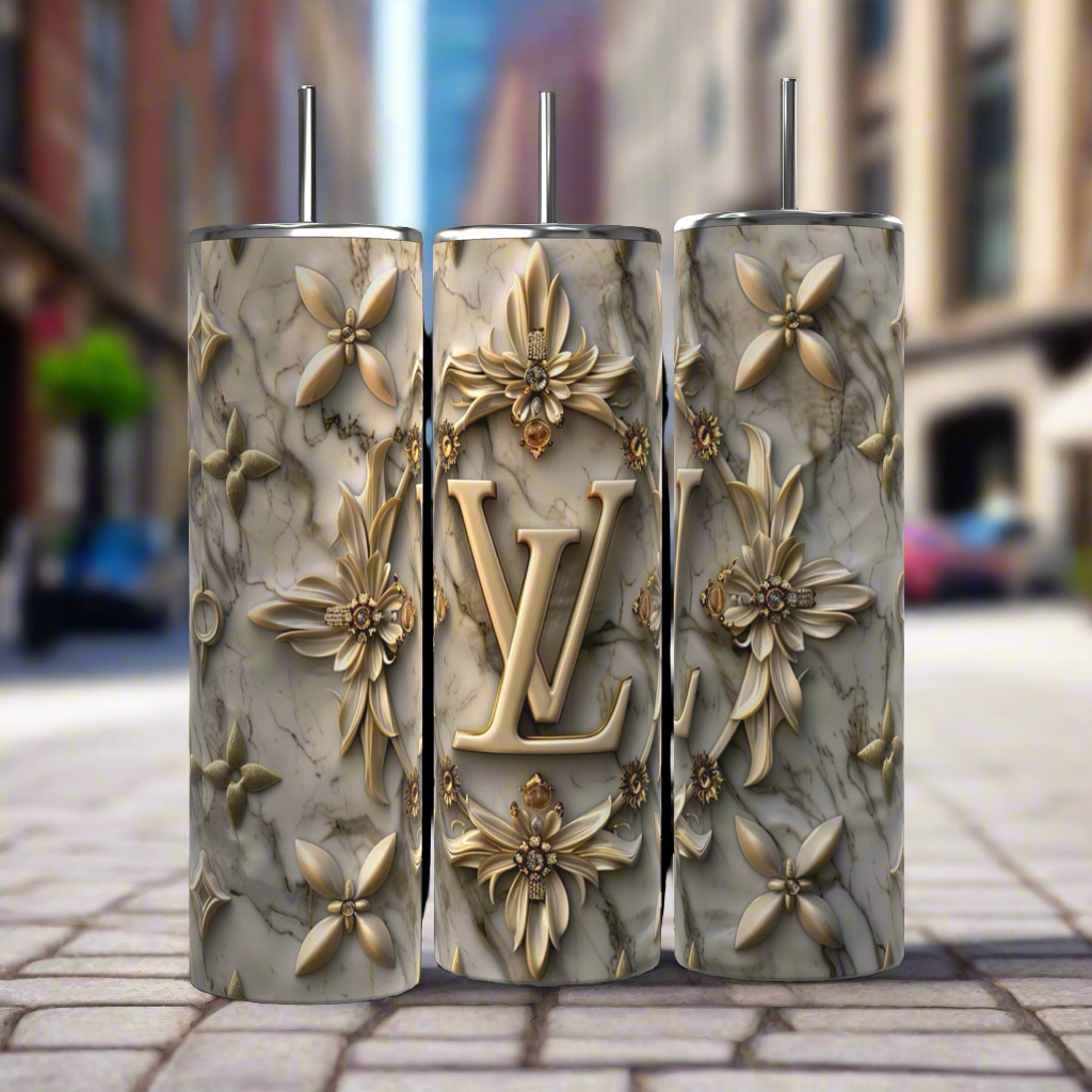 Three intricately decorated 20 oz Louis Vuitton Silver Gold tumblers by Kreative Kreationz, featuring a marbled texture and golden floral designs, stand side by side on a cobblestone street. The middle tumbler prominently showcases the initials "LV" and comes with a reusable straw. In the background, an urban setting with blurred buildings and greenery is visible.