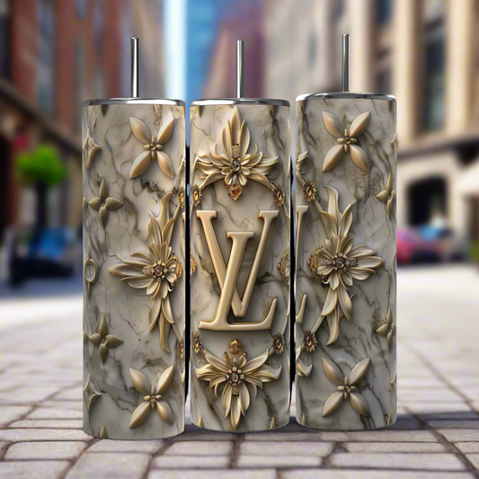 Three intricately decorated 20 oz Louis Vuitton Silver Gold tumblers by Kreative Kreationz, featuring a marbled texture and golden floral designs, stand side by side on a cobblestone street. The middle tumbler prominently showcases the initials "LV" and comes with a reusable straw. In the background, an urban setting with blurred buildings and greenery is visible.