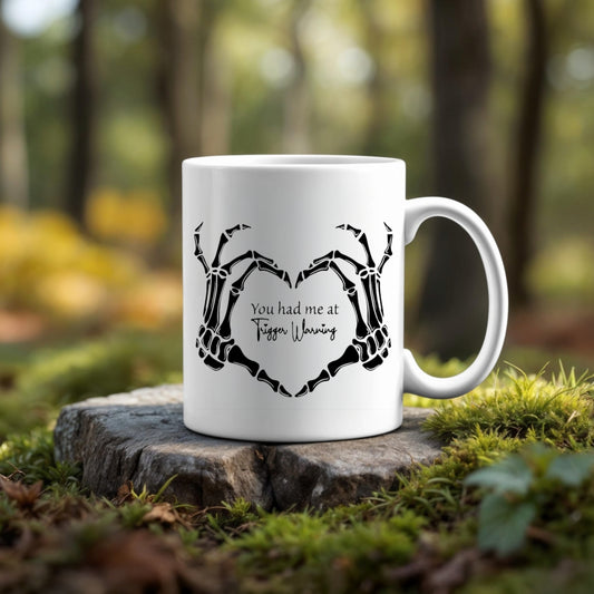 The Trigger Warning Mug by Kreative Kreationz, featuring skeleton hands forming a heart and "You had me at Tiffen Warning" text, rests on a tree stump against a blurred green forest backdrop—ideal for fans of unique photographic designs.