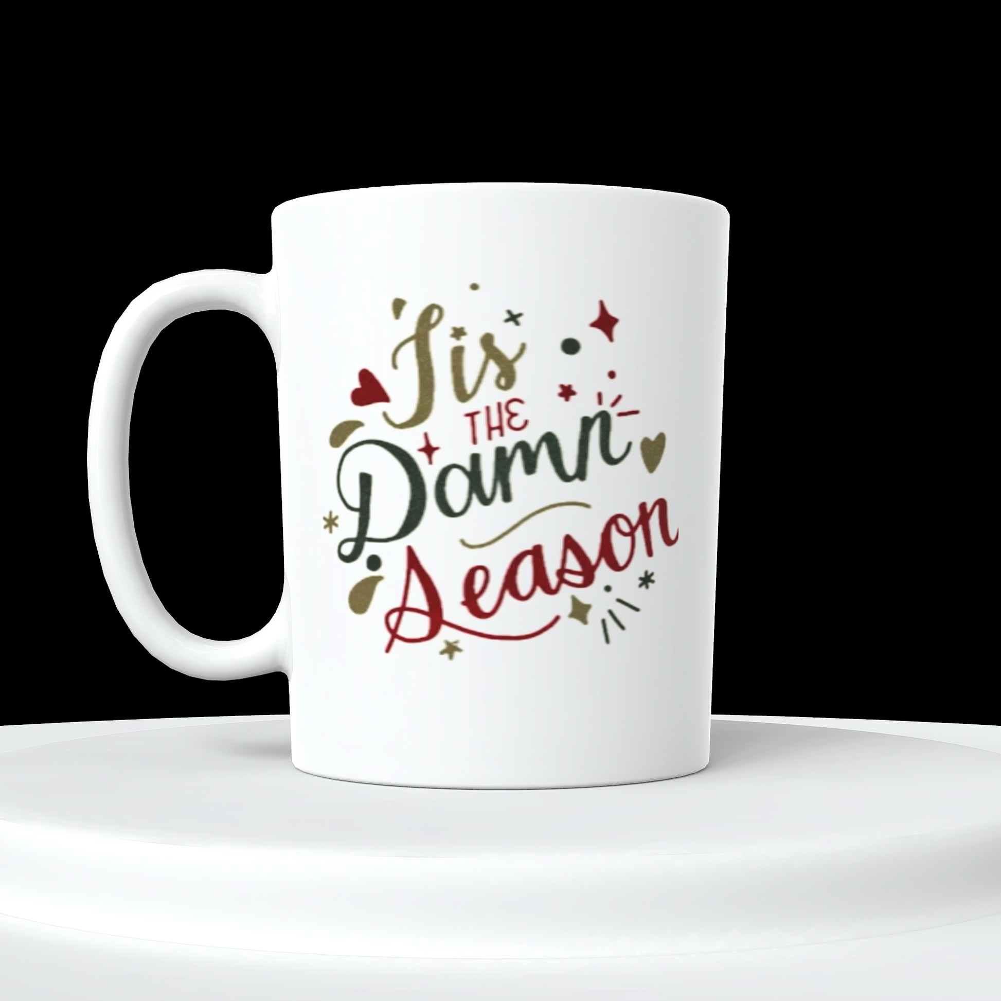 The Tis The Damn Season Taylor Swift Ceramic Mug by Kreative Kreationz features holiday-themed text reading "'Tis the Damn Season" in red and green decorative font, surrounded by small festive illustrations such as stars and hearts. The white ceramic mug is displayed on a white circular platform against a black background, making it an ideal gift for any Taylor Swift fan.