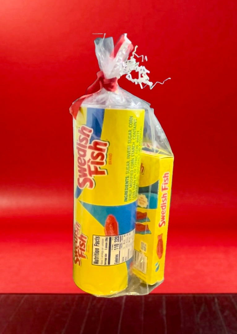 A plastic bag containing a Swedish Fish Gift Set with a 20oz stainless steel skinny tumbler, featuring yellow and blue packaging, branded "Swedish," with an illustration of a red fish. Next to it, a smaller box labeled "Swedish Fish" sits on the floor. The background is red with wooden flooring.