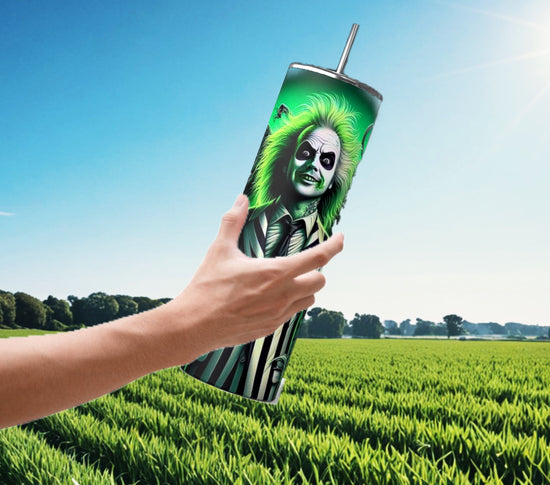 A hand holds the Beetlejuice Neon 20oz Tumbler by Kreative Kreationz, showcasing a vibrant design of a green-haired character in a striped suit. Set against a lush green field and blue sky, this drinkware is ideal for Halloween sips.