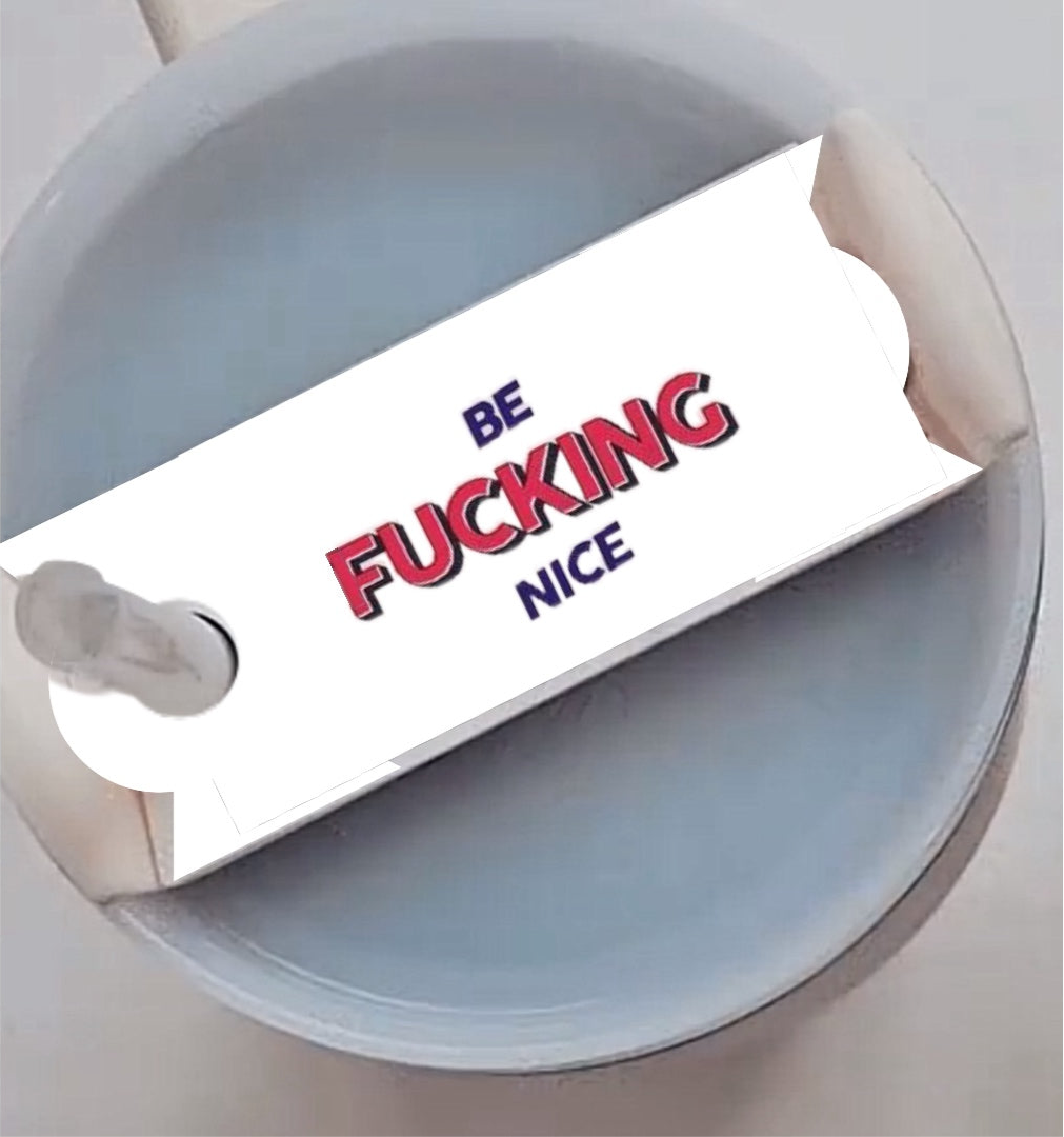 Close-up of a round, gray-colored object with a rectangular card attached to a stick in the middle. The card has the words "BE FUCKING NICE" printed on it in bold red and black lettering, functioning as a unique Stanley Lid Plate/Topper for your 20-ounce, 30-ounce, or 40-ounce Stanley cups from Kreative Kreationz.