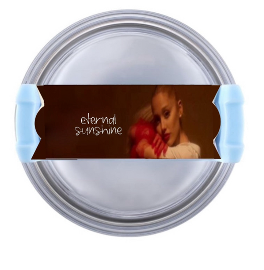 The Ariana Grande "External Sunshine" Stanley lid topper by Kreative Kreationz is a round object with a blue handle featuring a label of "eternal sunshine" and an image of someone holding a red object against a brown background, adding flair to your drinkware collection.