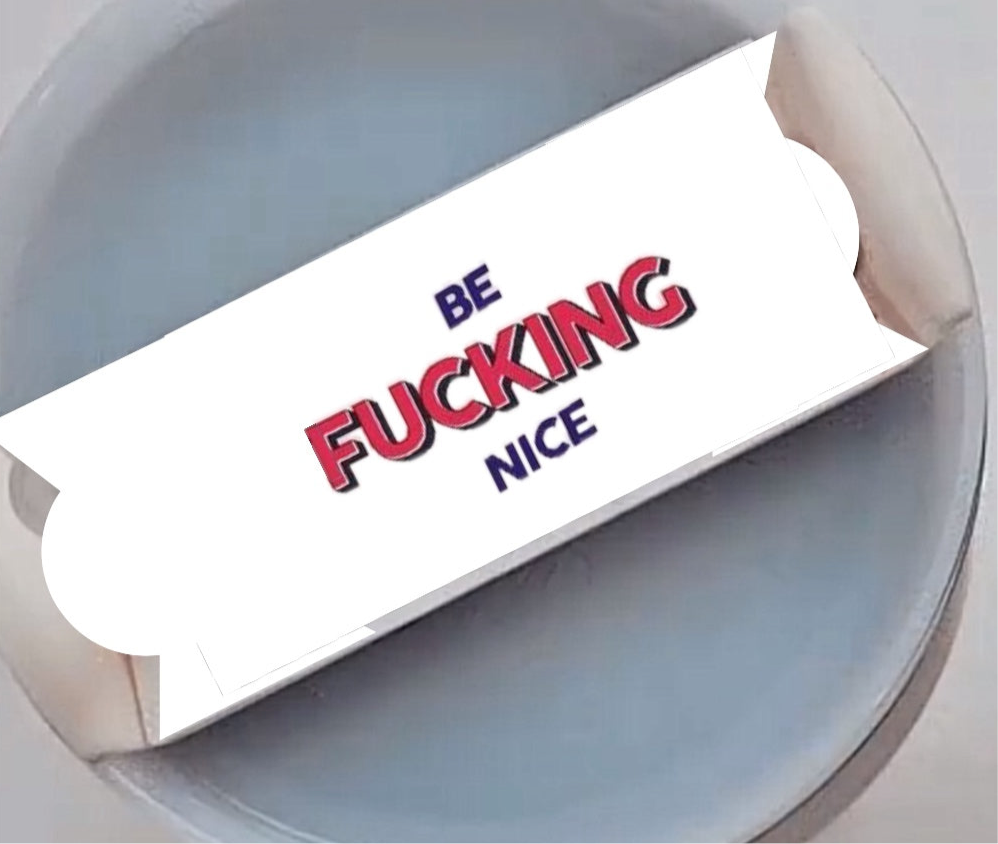 Close-up of a round, gray-colored object with a rectangular card attached to a stick in the middle. The card has the words "BE FUCKING NICE" printed on it in bold red and black lettering, functioning as a unique Stanley Lid Plate/Topper for your 20-ounce, 30-ounce, or 40-ounce Stanley cups from Kreative Kreationz.