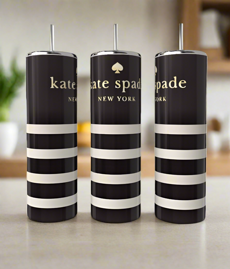 Three Kate Spade 20oz Skinny Tumblers from Kreative Kreationz, featuring black and white stripes with silver lids and straws, sit elegantly on a countertop. The blurred kitchen background, with plants and shelves, enhances the drinkware's allure.