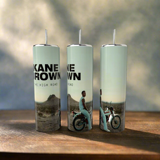 Three eco-friendly Kreative Kreationz "Kane Brown The High Road" 20oz Tumblers, depicting a desert scene with a motorcyclist, sit on a wooden table against a blurred background.