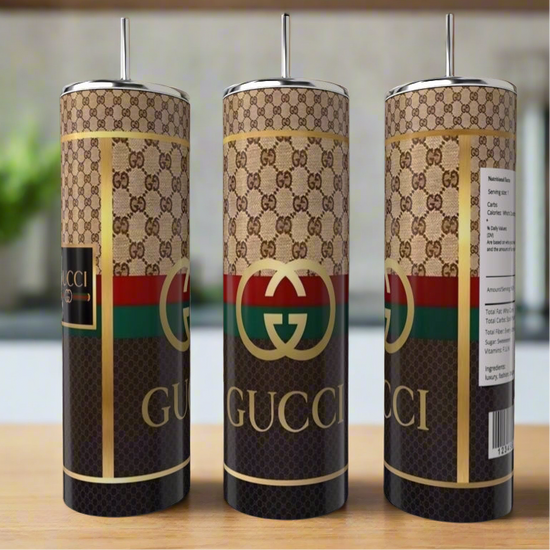 Three Gucci 20oz Skinny Tumblers by Kreative Kreationz, each with iconic GG patterns in brown, gold, red, and green, come with reusable straws. Displayed on a wooden surface against a blurred background, these tumblers feature double-wall insulation for optimal temperature control.