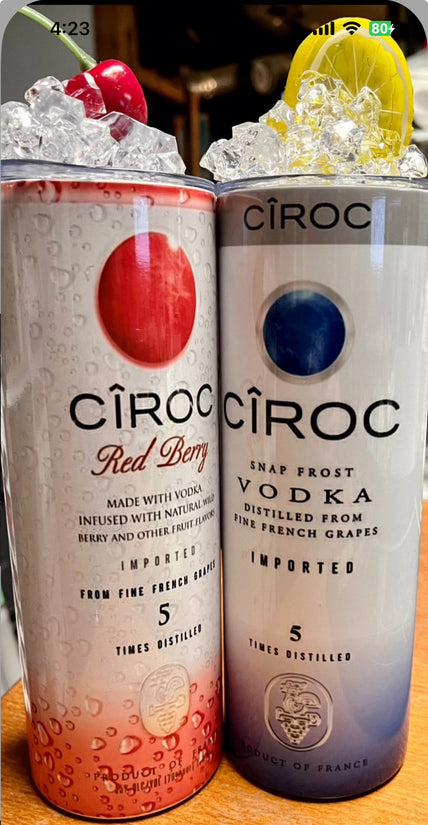 A lineup of CÎROC vodka cans in various flavors including original, mango, red berry, coconut, watermelon, and peach is beautifully arranged on an outdoor surface with a green hedge and blue sky background. Each can is paired with a chic 20oz Ciroc Tumbler with 3D Lid Topper by Kreative Kreationz.