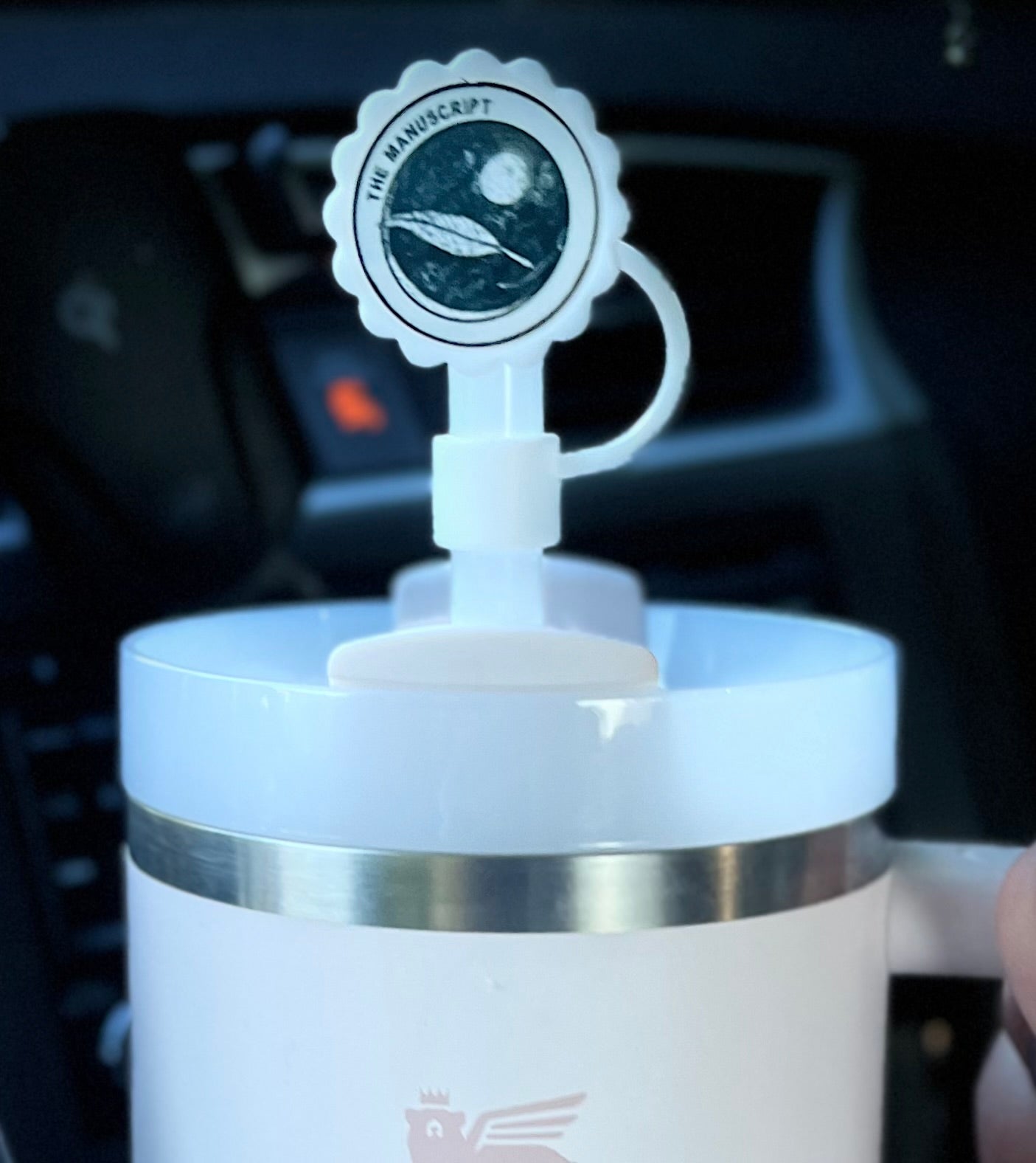 A white cup with a handle displays a round sticker on the lid, which features an illustration of a planet and spacecraft, accompanied by the text "THE MANUSCRIPT" around it. The background is black and the image appears in black and white. For added convenience, it pairs well with Kreative Kreationz's Straw Cover Topper, which fits Stanley cup sizes 40oz, 30oz, and 20oz with 10mm straws.