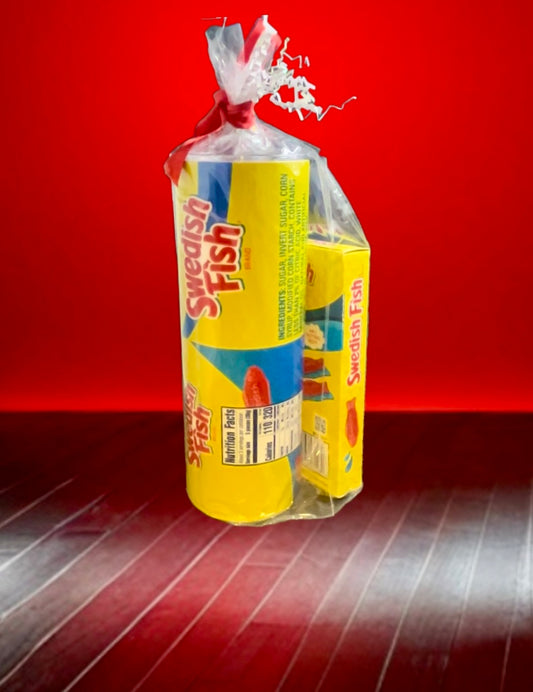 A plastic bag containing a Swedish Fish Gift Set with a 20oz stainless steel skinny tumbler, featuring yellow and blue packaging, branded "Swedish," with an illustration of a red fish. Next to it, a smaller box labeled "Swedish Fish" sits on the floor. The background is red with wooden flooring.