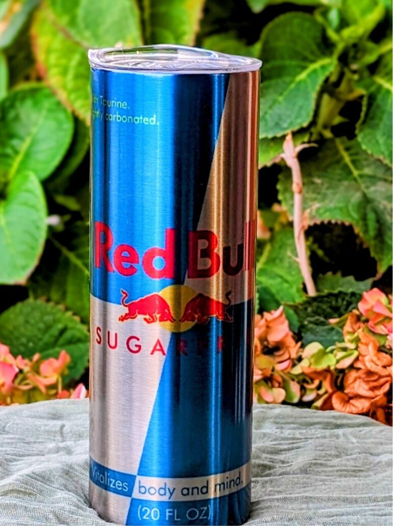 A tall Red Bull Sugar Free Silver Stainless Steel 20oz Tumbler by Kreative Kreationz rests on a gray surface, surrounded by vibrant green leaves and a few flowers. Its sleek silver design mirrors the eye-catching presence of an energy drink, ready to energize with its striking appearance.