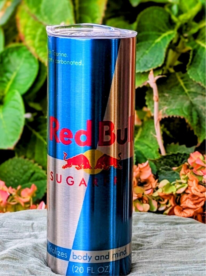 A 20 oz RB Energy-Silver Stainless Steel Tumbler from Kreative Kreationz, resembling a Red Bull Sugar Free can, stands on fabric with leafy greens and pink flowers in the background. The silver and blue tumbler features the Red Bull design, highlighting its revitalizing benefits.