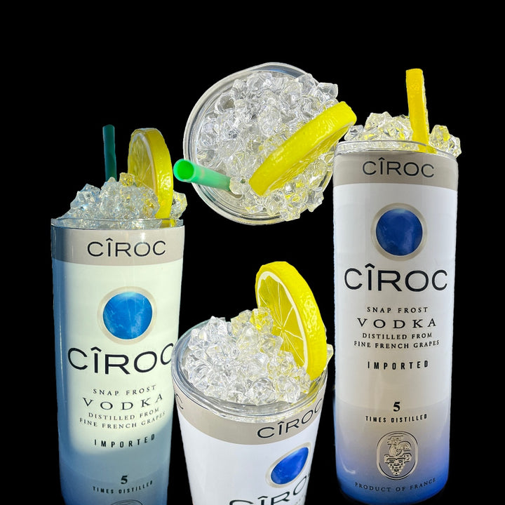 Two cans of Kreative Kreationz Ciroc 20oz Tumbler with 3D Lid Topper are shown on a wooden surface, reminiscent of travel tumblers. The left can is Red Berry flavor, topped with ice and a cherry. The right can is Vodka Snap Frost flavor, topped with ice and a lemon slice. Both cans feature product information and branding.