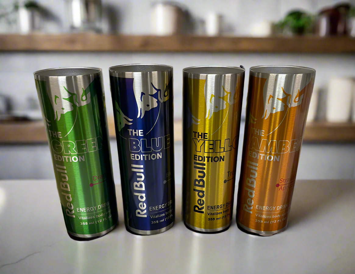 Four colorful cans of Red Bull energy drinks are lined up on a counter. From left to right, the cans are green (Green Edition, Dragon Fruit flavor), blue (Blue Edition, Blueberry flavor), yellow (Yellow Edition, Tropical flavor), and orange (Amber Edition, Strawberry-Apricot flavor). Next to them sits a stylish Red Bull Stainless Steel 20oz Tumbler by Kreative Kreationz in a chic kitchen setting.