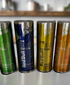 Four colorful cans of Red Bull energy drinks are lined up on a counter. From left to right, the cans are green (Green Edition, Dragon Fruit flavor), blue (Blue Edition, Blueberry flavor), yellow (Yellow Edition, Tropical flavor), and orange (Amber Edition, Strawberry-Apricot flavor). Next to them sits a stylish Red Bull Stainless Steel 20oz Tumbler by Kreative Kreationz in a chic kitchen setting.