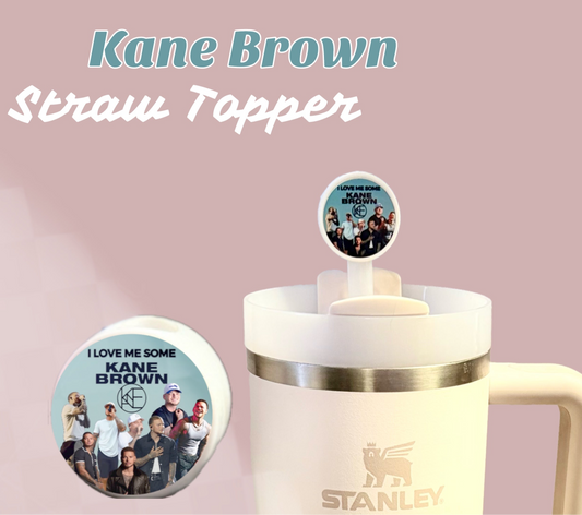 A Kreative Kreationz's Kane Brown Straw Topper, featuring a circular image of people and "I love me some Kane Brown" text, adorns a beige Stanley tumbler with a metal rim and handle.