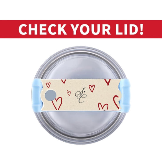 A silver tumbler filled with black liquid features a beige piece of tape adorned with red hearts and a cursive letter 